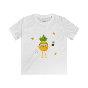 Op de afbeelding: Wit T-shirt met een cartoon ananas karakter met een bril en een beker en een drankje. De ananas heeft gele benen en armen. Gouden sterren zijn verspreid rond de ananas. Het shirt is gemaakt van een zacht materiaal.