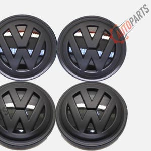 Könnte beinhalten: Vier schwarze Radnabenabdeckungen mit dem Volkswagen-Logo. Die Kappen sind kreisförmig mit einem erhabenen schwarzen VW-Emblem in der Mitte. Der Hintergrund ist weiß.