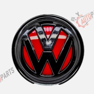 Peut inclure: Un emblème circulaire Volkswagen présentant une finition noire brillante sur les lettres V et W en relief, sur un fond rouge vif. L'emblème possède une surface lisse et réfléchissante avec une bordure noire circulaire, et le texte AUTOPARTS est visible.