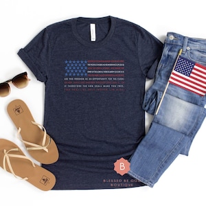 Könnte beinhalten: Ein dunkelblaues Kurzarm-T-Shirt mit einer Grafik der amerikanischen Flagge aus Text, dazu eine blaue Jeans, beige Flip-Flops und braun getönte Sonnenbrillen. Eine kleine amerikanische Flagge an einem Stab steckt in der Hosentasche.