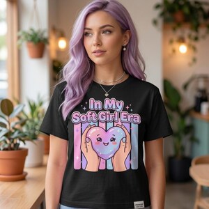Pode incluir: Camiseta preta com a frase "In My Soft Girl Era" e um desenho de um coração segurado por duas mãos. O coração é rosa, azul e roxo com um rosto sorridente. Listras verticais rosa, azul e roxo estão atrás do coração.