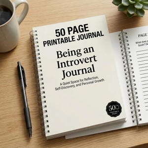 Könnte beinhalten: Ein weißes Spiralbuch mit dem Titel "Being an Introvert Journal" und dem Text "50 Page Printable Journal". Ein schwarzer Stift und eine Kaffeetasse liegen auf einer Holzoberfläche. Das Notizbuch ist auf einer linierten Seite geöffnet.