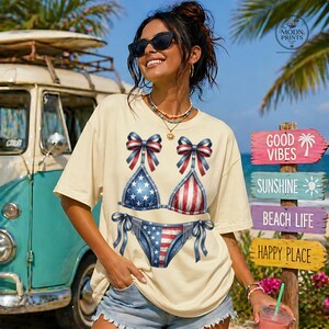 Peut inclure: T-shirt crème avec un motif de bikini patriotique aux étoiles et rayures rouges, blanches et bleues. Le t-shirt a une coupe décontractée et des manches courtes. L'image comprend également un van vintage et une signalisation sur le thème de la plage.