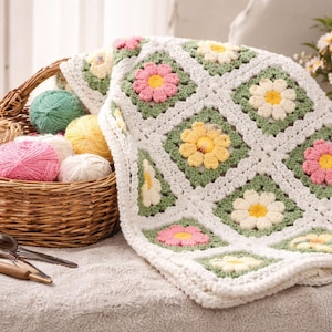 May include: A handmade crochet blanket with a daisy pattern in pink, yellow, and white, resting on a table. A wicker basket filled with yarn balls in various colors sits nearby, along with crafting tools.