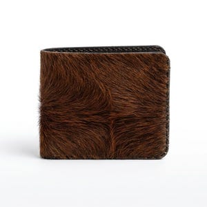 Puede incluir: Una cartera plegable con un exterior marrón texturizado de piel de animal. La cartera tiene un interior y bordes de cuero marrón oscuro o negro. La cartera es rectangular y parece nueva.