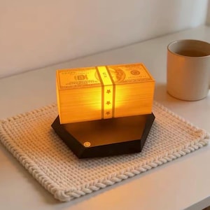 May include: A decorative lamp shaped like a stack of $100 bills, illuminated with a warm glow. The lamp is secured with a band featuring star accents and rests on a black, hexagonal base. It sits on a cream-colored, knitted coaster.