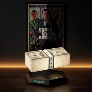 Op de afbeelding: Een stapel Amerikaanse dollarbiljetten, vastgezet met een witte band en groene sterren, zwevend boven een zwarte basis. Op de basis staat het woord "LEVITATIC". Op de achtergrond een ingelijste poster van Grand Theft Auto 5.