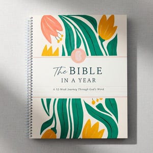 May include: A spiral-bound book titled "The Bible in a Year" with a floral design. The cover features a white background with green leaves, orange and yellow flowers, and the text "The Bible in a Year" in a script font.
