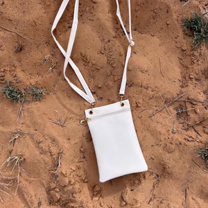 May include: A white crossbody bag with a textured surface and a long, white strap. The bag has a zippered top and gold-colored hardware. The strap is attached to the bag with gold-colored rings.