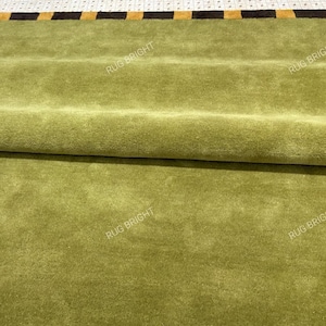 May include: A solid olive green area rug with a thick border featuring alternating black and mustard yellow rectangular blocks. The rug has a soft, dense pile texture and is partially folded over to reveal the underside and the edge detail.