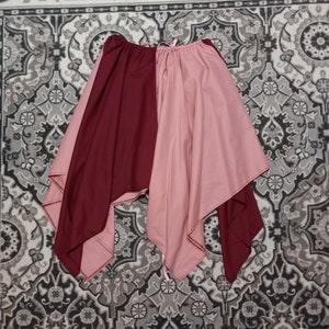 May include: A two-toned, half-and-half skirt in burgundy and blush pink. The skirt has a gathered waistband and a flowing, asymmetrical hemline. The fabric appears to be a soft, lightweight material, suitable for layering or wearing on its own.