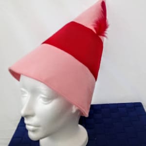 Puede incluir: Un sombrero cónico rosa y rojo con una pluma roja. El sombrero se exhibe en una cabeza de maniquí blanca. El sombrero tiene un borde rosa y una banda roja en el medio.