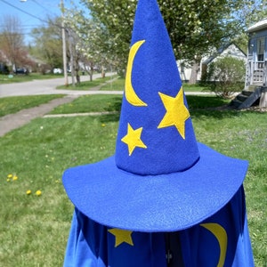 Puede incluir: Un sombrero puntiagudo azul brillante y una capa a juego con aplicaciones de fieltro amarillo en forma de lunas crecientes y estrellas de cinco puntas. El sombrero tiene un ala ancha y una copa cónica alta, mientras que la capa cae hacia abajo.
