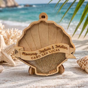 May include: Wooden seashell ornament with a banner that reads "Mommy, Joshua & Jordan CABO SAN LUCAS 2026". The ornament is light brown and has a small loop at the top for hanging. The background shows a beach scene with seashells.