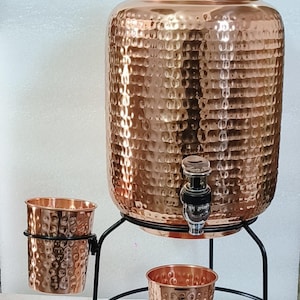 May include: A large, hammered copper water dispenser with a matching lid and a metal spigot. The dispenser sits on a black metal stand that includes a side holder for a hammered copper cup, with an additional matching cup placed below the spigot.