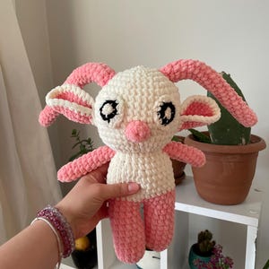 May include: A handmade crochet doll in white and pink. The doll has large, black eyes, a pink nose, and pink ears, arms, and legs. The doll is being held in a person's hand. The doll is a cute, whimsical design.