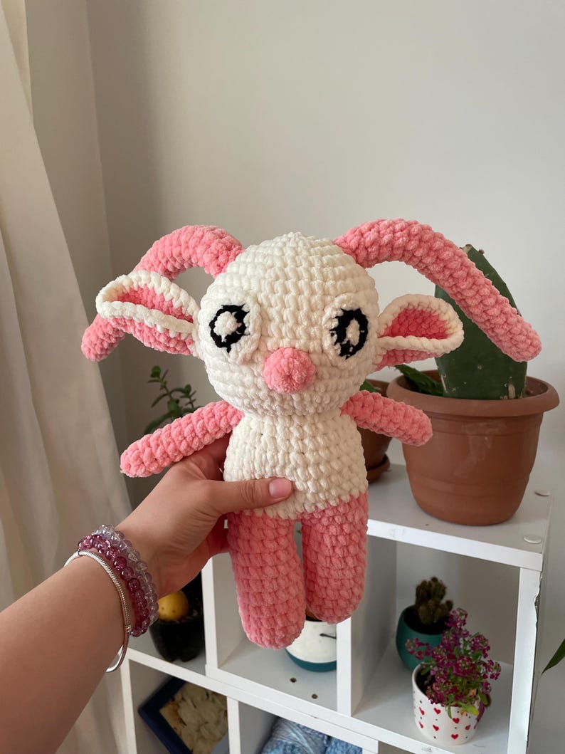 May include: A handmade crochet doll in white and pink. The doll has large, black eyes, a pink nose, and pink ears, arms, and legs. The doll is being held in a person's hand. The doll is a cute, whimsical design.