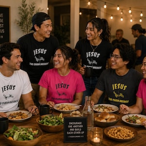 May include: A group of people are seated around a wooden table, smiling and eating. They are wearing t-shirts with the word "JESUS" and the phrase "THE CHAIN BREAKER". A sign reads "WE LOVE BECAUSE HE FIRST LOVED US".