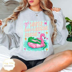 May include: A light grey sweatshirt with a graphic design. The design features a pink flamingo float with a green alligator, the text "HOTTER THAN A HOOCHIE COOCHIE", and stars. The sweatshirt has a crew neck and long sleeves.