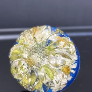 Puede incluir: Un pisapapeles circular de resina que presenta una colección de flores de margarita blancas secas con centros amarillos incrustadas en un material transparente y brillante. La base del objeto es de un color azul profundo sólido y contrastante.