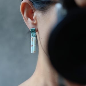 May include: A drop earring featuring a rectangular top section made of small, woven turquoise beads. Attached below are two vertical, pale blue cylindrical beads with subtle orange-brown speckles, creating a linear, multi-textured design.