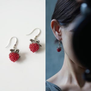 May include: A pair of dangle earrings featuring small, three-dimensional strawberry charms crafted from tiny red beads. The berries are topped with textured, metallic silver-toned leaves and attached to simple silver-toned ear wires for hanging.