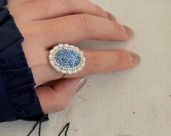 May include: A ring featuring an oval centrepiece composed of small, light blue square tiles, bordered by a ring of tiny metallic gold beads and an outer edge of small white seed pearls, set on a thin, simple gold-toned metal band.