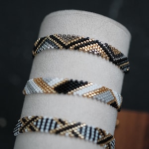 May include: Three beaded bracelets featuring intricate geometric patterns. The top bracelet uses black, gold, and silver beads. The middle bracelet displays white, gold, black, and gray beads. The bottom bracelet features a white, black, and gold design.