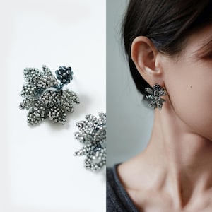 May include: A pair of stud earrings crafted from small, metallic silver and dark blue beads arranged in a floral shape. The design features a cluster of textured, shimmering petals with a single dark blue bead at the center of each earring.