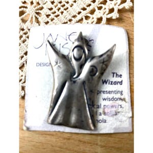 May include: A silver-toned metal wizard pin with outstretched wings, set on a white card with the text "The Wizard presenting wisdom". The pin is from "Jane Nis Design". The background is a wooden surface with a lace doily.
