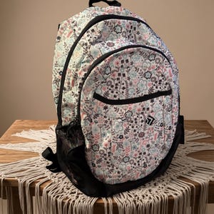 May include: A floral-patterned backpack with a white background and black accents. The design features pink, blue, and gray flowers and foliage. The backpack has a front pocket, side mesh pocket, and a top handle.