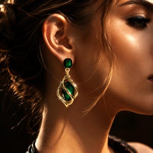 Puede incluir: Un pendiente colgante de tono dorado con una piedra preciosa verde esmeralda facetada en el poste y una piedra ovalada verde esmeralda más grande encerrada en una estructura de metal dorado en forma de jaula que cuelga.
