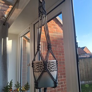 May include: A grey macrame plant hanger holding a white and blue ceramic pot with a black dot pattern. The hanger is knotted and has a tassel. The pot is suspended in front of a window, with a brick wall visible.