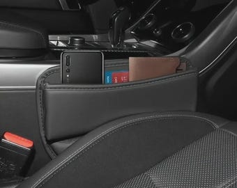 Pode incluir: Organizador de assento de carro preto com vários compartimentos. Um smartphone, carteira e cartões são visíveis no interior. O organizador é feito de um material escuro e liso e está posicionado entre os bancos do carro.