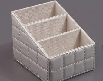 Pode incluir: Um organizador de mesa prateado com três compartimentos. O exterior apresenta um padrão acolchoado com costuras brancas. O interior é de tecido bege claro. O organizador é inclinado para facilitar o acesso.
