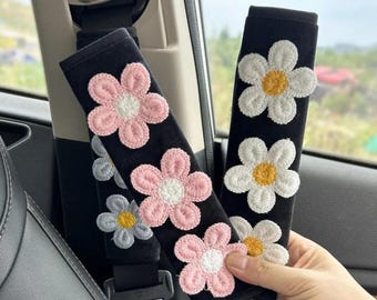 Pode incluir: Capas pretas para cintos de segurança com desenhos de flores bordadas. Uma capa apresenta flores rosa e brancas, outra tem flores brancas e amarelas. As capas são projetadas para caber nas correias do cinto de segurança de um carro.