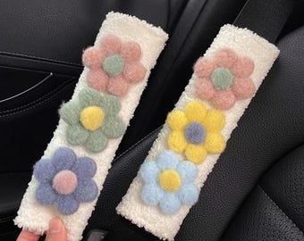 Pode incluir: Duas capas brancas e macias para cintos de segurança adornadas com flores de feltro coloridas. Cada capa apresenta três flores em tons de rosa, amarelo, azul e verde. As capas são projetadas para serem presas a um cinto de segurança do carro.