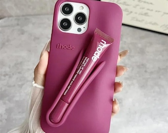 Pode incluir: Uma capa de telefone magenta com o nome da marca "rhode" em relevo na parte de trás. Um tubo de bálsamo labial magenta a condizer com a palavra "mode" e texto está preso à capa. A capa do telefone tem uma pega curva.
