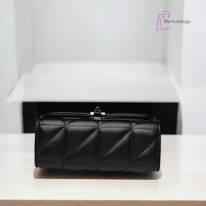 Puede incluir: Un bolso rectangular negro acolchado con un cierre plateado. El bolso tiene un patr&oacute;n de diamantes. El bolso est&aacute; hecho de un material suave y est&aacute; sobre una superficie blanca.