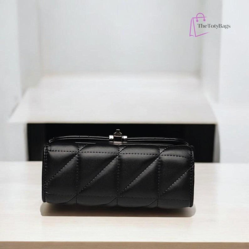 Puede incluir: Un bolso rectangular negro acolchado con un cierre plateado. El bolso tiene un patr&oacute;n de diamantes. El bolso est&aacute; hecho de un material suave y est&aacute; sobre una superficie blanca.