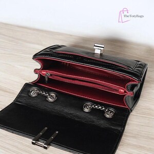 Puede incluir: Bolso negro acolchado con interior rojo. El bolso tiene un cierre plateado y correas de cadena. El interior tiene un bolsillo con cremallera. El bolso est&aacute; abierto, revelando el interior. La marca "The Toty Bags" est&aacute; en la esquina superior derecha.