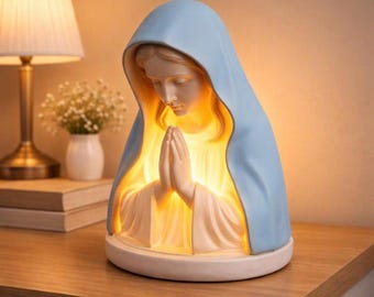 Pode incluir: Uma estatueta religiosa da Virgem Maria com um véu azul claro e um interior quente e brilhante. A estátua apresenta as mãos juntas em oração e está sobre uma base branca. A lâmpada está sobre uma superfície de madeira.