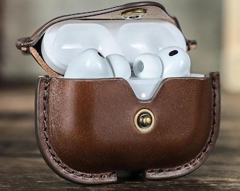 Pode incluir: Uma capa de couro castanho com fecho de pressão, contendo fones de ouvido sem fio brancos. A capa está aberta, revelando os fones de ouvido. A capa tem uma borda costurada e um botão de pressão cor de latão.