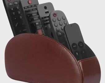Pode incluir: Um suporte de couro castanho para controle remoto com vários controles remotos pretos. O suporte é semicircular com uma borda costurada. Os controles remotos têm vários botões e etiquetas, incluindo "INPUT" e números.