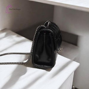 Puede incluir: Bolso de hombro negro acolchado con cadena plateada. El bolso tiene forma rectangular con cierre de solapa y cierre plateado. Fabricado con un material brillante y liso.