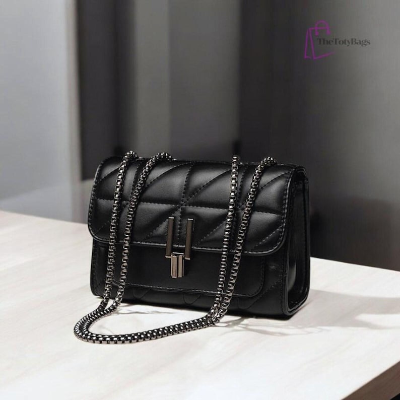 Puede incluir: Bolso bandolera negro acolchado con cadena plateada. El bolso tiene forma rectangular con cierre de solapa y cierre met&aacute;lico plateado. El logotipo de TotyBags est&aacute; en la esquina superior derecha.