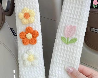 Pode incluir: Duas capas brancas para cintos de segurança com textura de waffle. Uma tem três flores bordadas em amarelo, laranja e branco. A outra tem uma tulipa rosa com folhas verdes. Acessórios para interiores de automóveis.