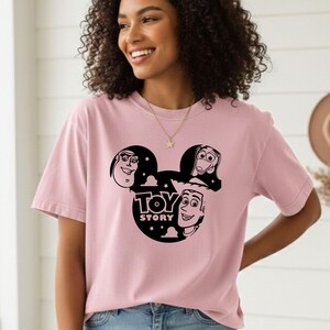 K&ouml;nnte beinhalten: Hellrosa T-Shirt mit schwarzer Grafik von Toy Story-Charakteren in einer Mickey Mouse-Silhouette. Der Text "TOY STORY" ist ebenfalls schwarz. Das T-Shirt hat einen l&auml;ssigen Stil.