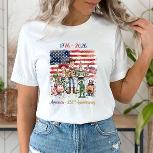 May include: White t-shirt featuring a graphic of the American flag with the text "1776-2026" and various Toy Story characters. The design celebrates America's 250th anniversary.