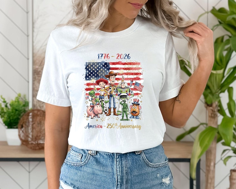 May include: White t-shirt featuring a graphic of the American flag with the text "1776-2026" and various Toy Story characters. The design celebrates America's 250th anniversary.